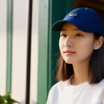 라코스테 클래식 캡과 프레드페리 테니스 캡 비교 - A young woman, in her early twenties, stands casually in a sunlit outdoor cafe. She is wearing a cla...